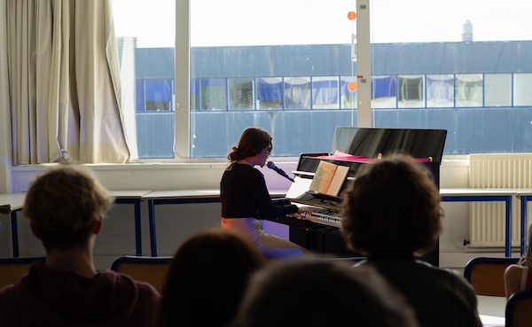 Une pianiste joue devant un public. En arrière-plan on aperçoit par la fenêtre le haut du bâtiment bleu de la faculté.